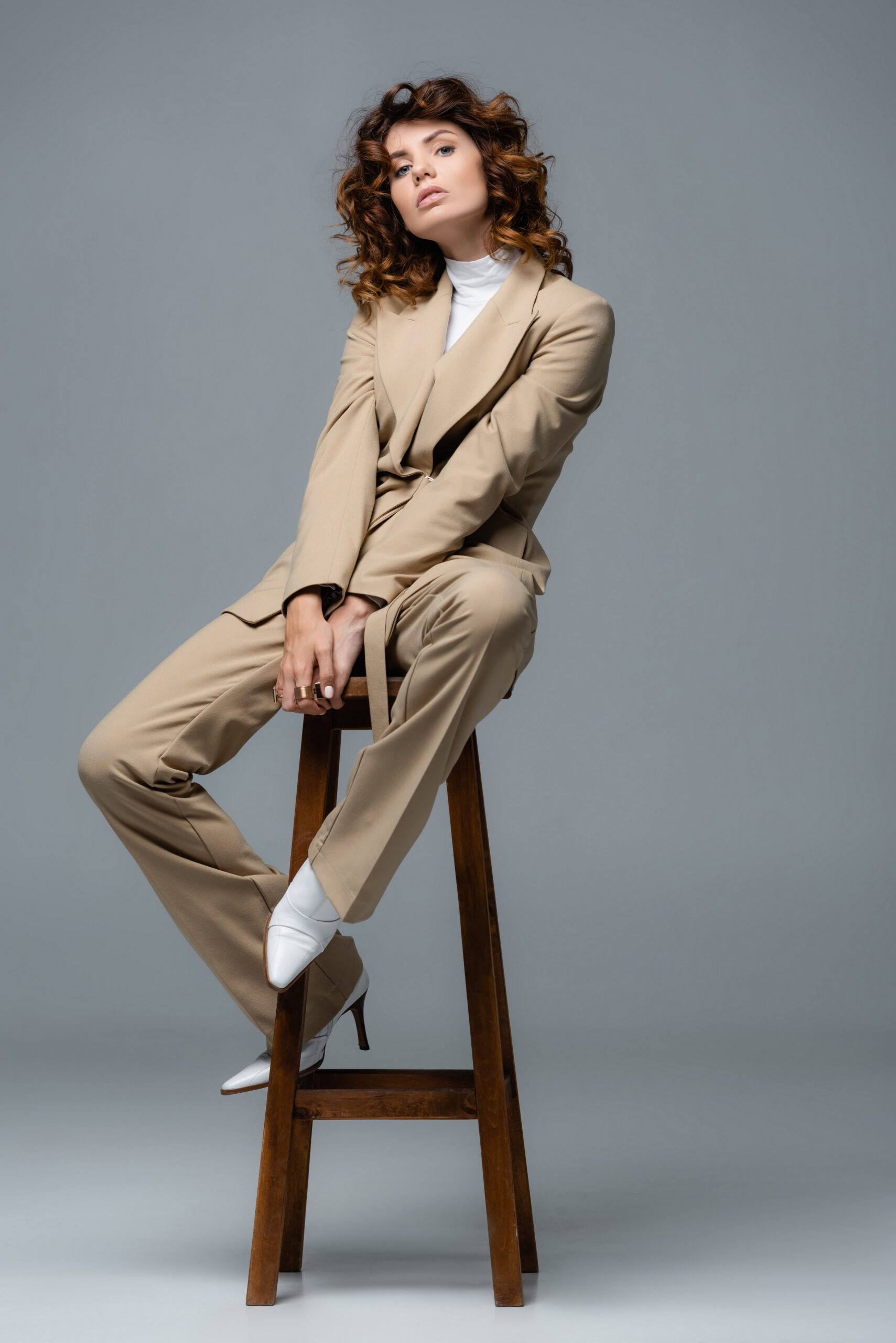 elegant woman in beige suit posing on wooden chair on grey background